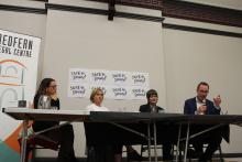 Three women and a man sit at a table up on a stage delivering a presentation in from of signs that say "Safe and Sound"