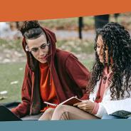 two students sit in grass with a laptop computer and book