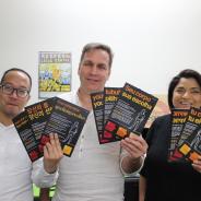 Three people stand holding multi-language fliers