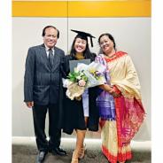 Three people stand smiling teh woman in the middle wears a graduation gown and motor board hat