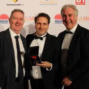Three men in suits stand in a row hold a medal in a black box
