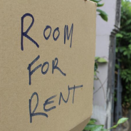 Text reads "Room for Rent" on piece of cardboard attached to fence on suburban street