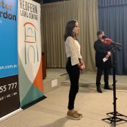 Young woman standing at microphone in front of two colourful banners 