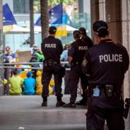 Group of police in dark blue uniform standing inside with backs to camera watching a group of people protesting outside 
